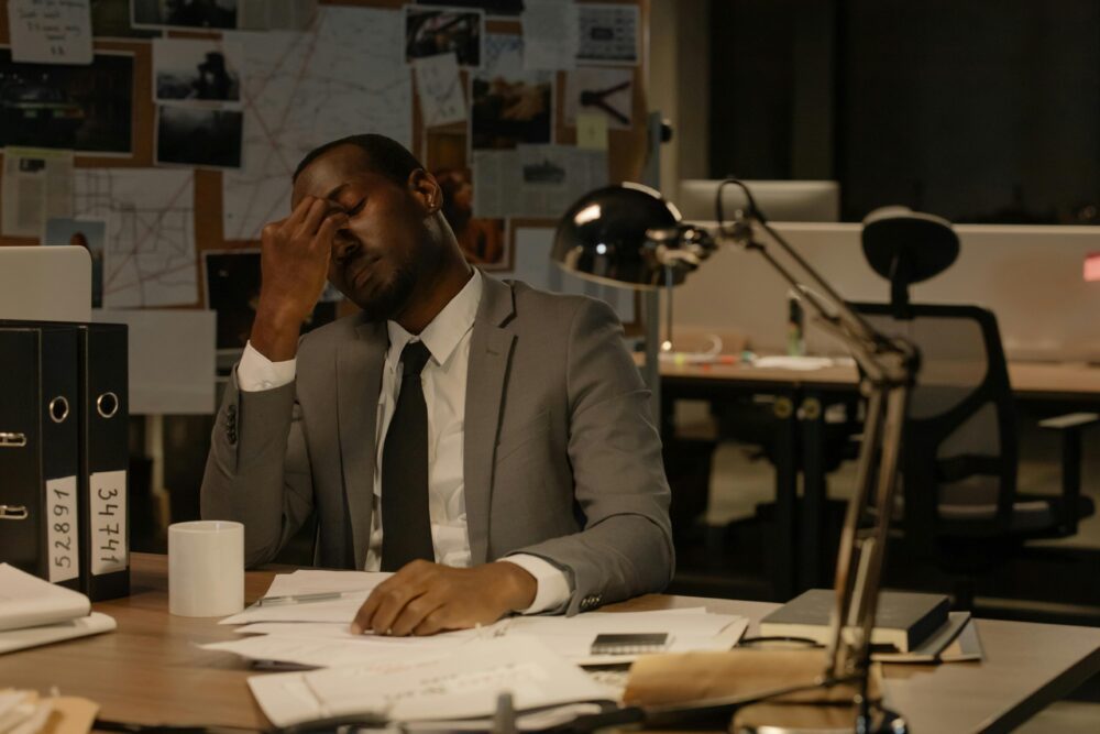 A tired employee in a suit sitting at his desk with his hand on his face, overwhelmed by paperwork in a dimly lit office